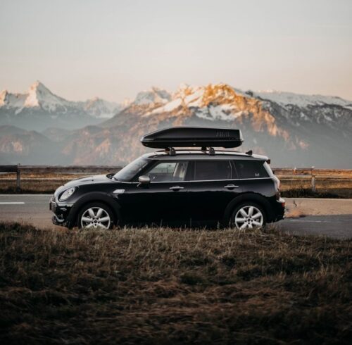 Mini noire équipée d'un coffre de toit pour un long voyage, qui roule sur route de montagne